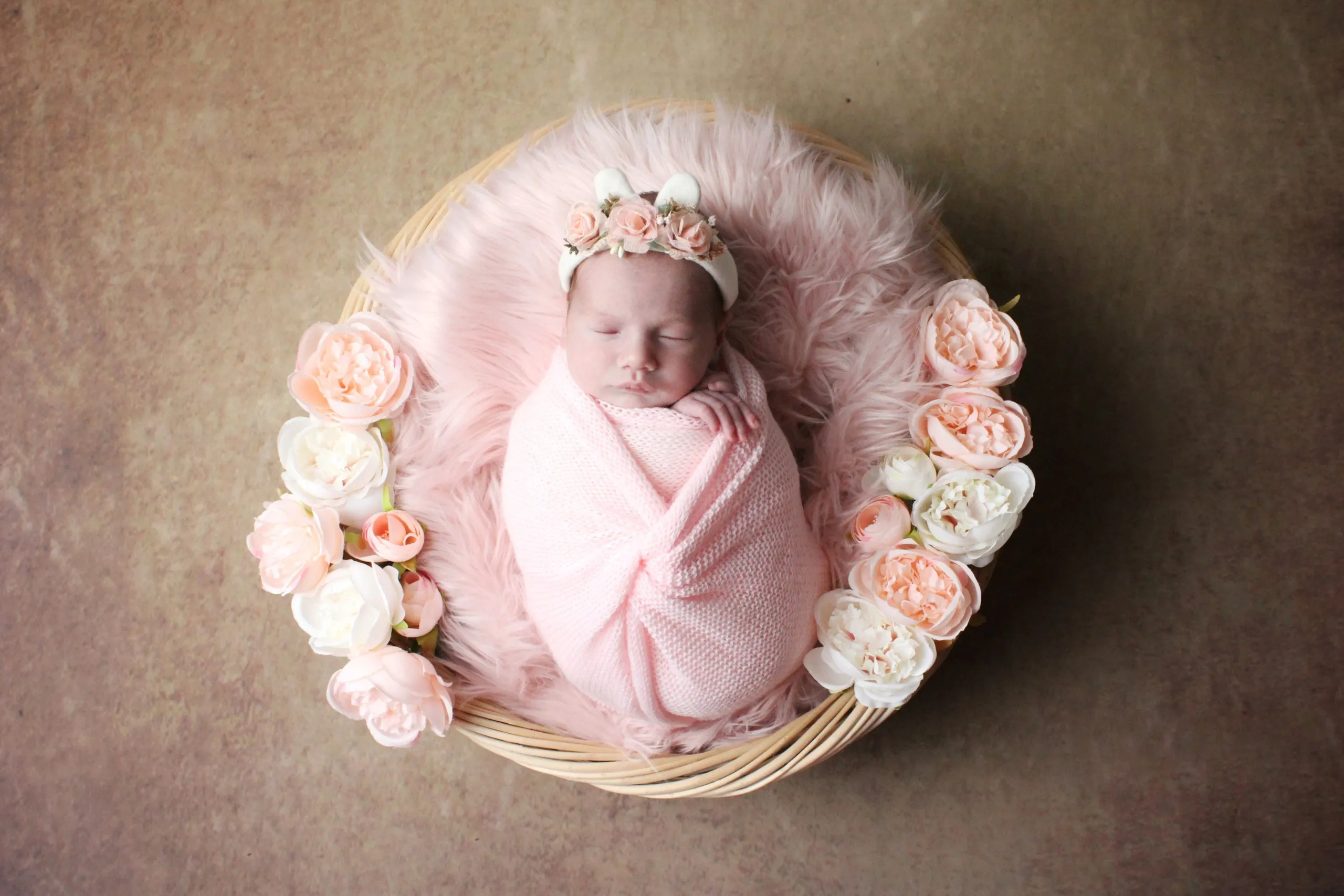 Neonata addormentata in un cesto di vimini, avvolta in una fascia di maglia rosa, con una coroncina di fiori e una ghirlanda di rose chiare attorno al cesto